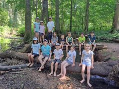 13 barfüßige Kinder der Wandergruppe sitzen auf einem Baumstamm und einer Brücke quer über dem Bach